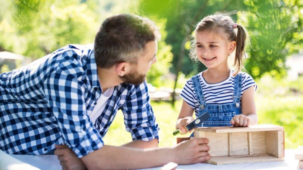 Father watching his young, smiling daughter build a wooden project outdoors.