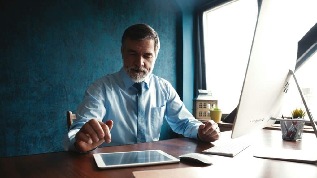 Older man in a dress shirt and tie sitting at a desk looking at a tablet.