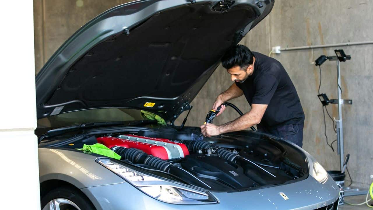Man in a black shirt cleaning the engine of a silver car with a hose.
