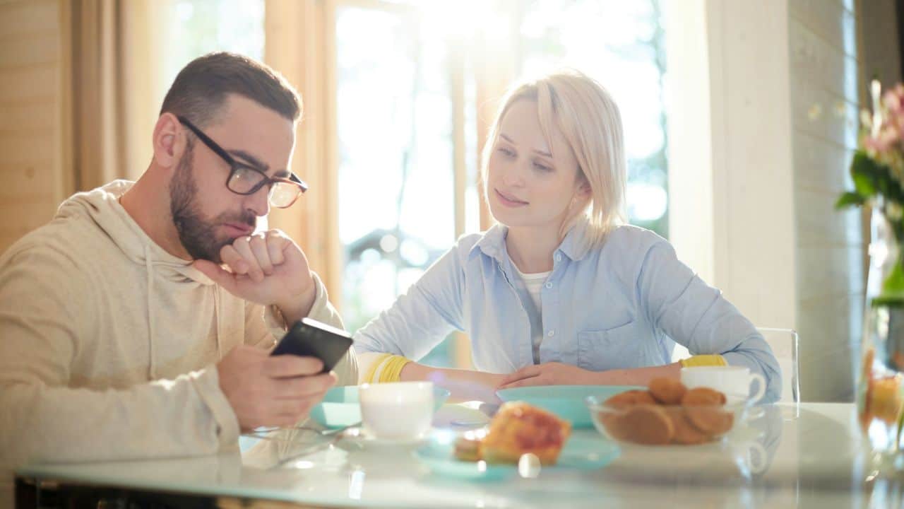 Man with beard and glasses looks at his phone while a woman smiles beside him at a table.