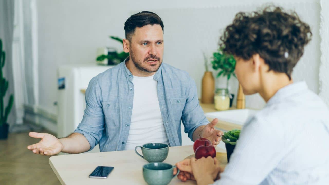 Man gesturing with open hands while talking to a woman across a kitchen table.
