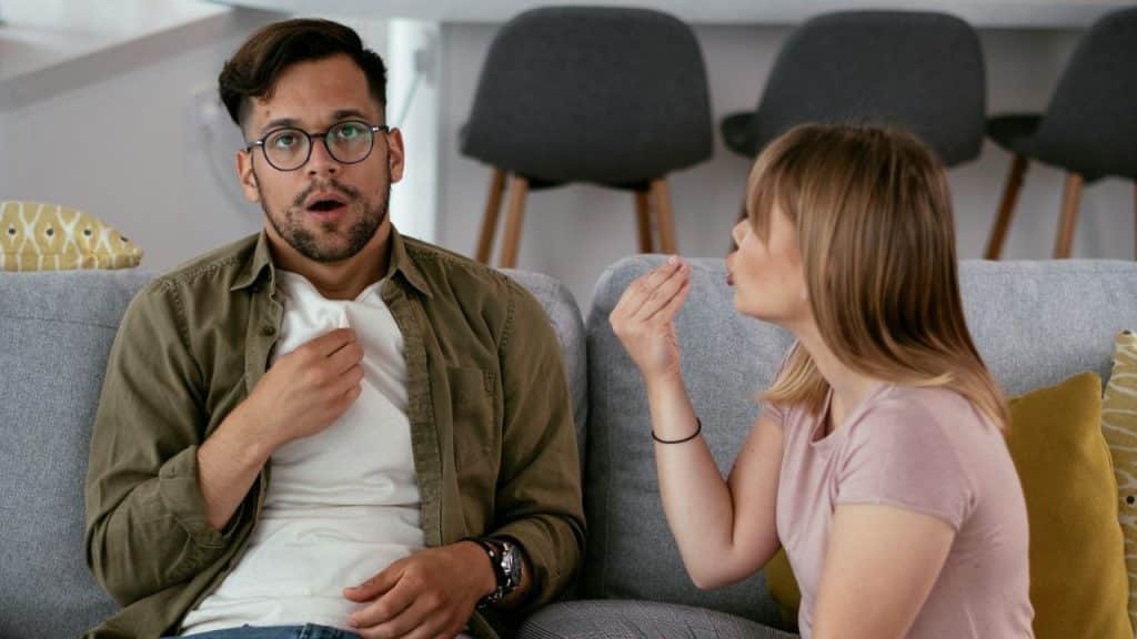 Man looking shocked with a hand on his chest next to a woman speaking animatedly.