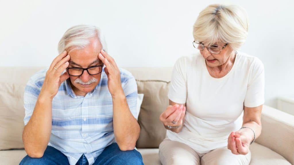 Older man sits with his head in his hands next to a woman holding a small object.