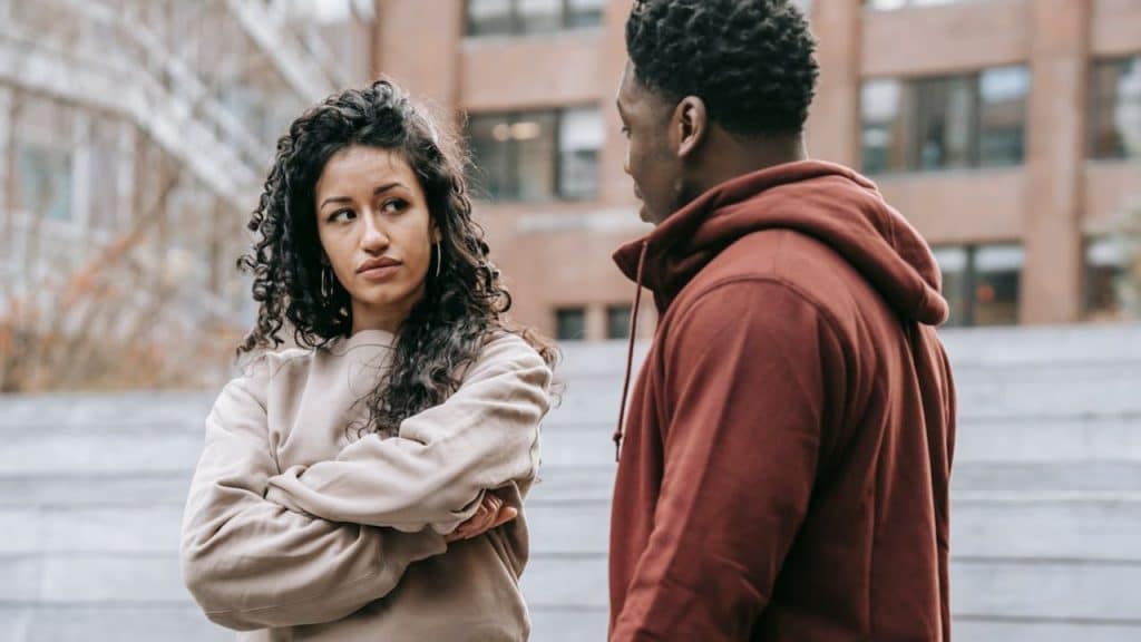 Woman with long curly hair and crossed arms looking suspiciously at a man outdoors.