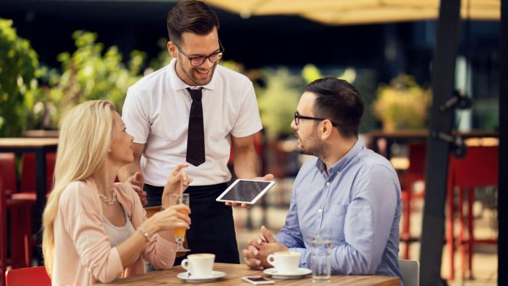 Smiling waiter holding a tablet while talking to a man and woman at an outdoor cafe.