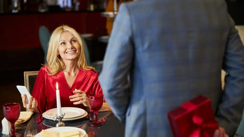 Smiling woman seated at a restaurant table while a man holds a red gift box behind his back.