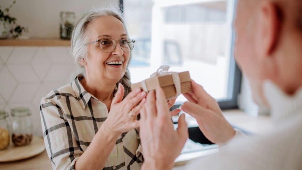A smiling older woman accepts a small, wrapped gift from a person whose back is to the camera.