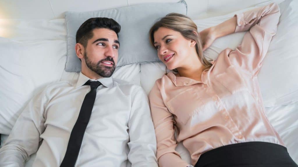 A man in a shirt and tie and a woman in a silk blouse lying on a bed looking at each other.
