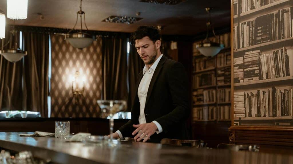 A man in a suit standing at a bar with drinks in front of him.