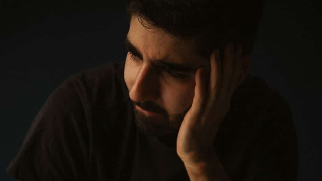 A bearded man looking sad and holding his palm up to his face.