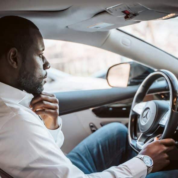 A man sitting in a car, holding the steering wheel and adjusting his collar.