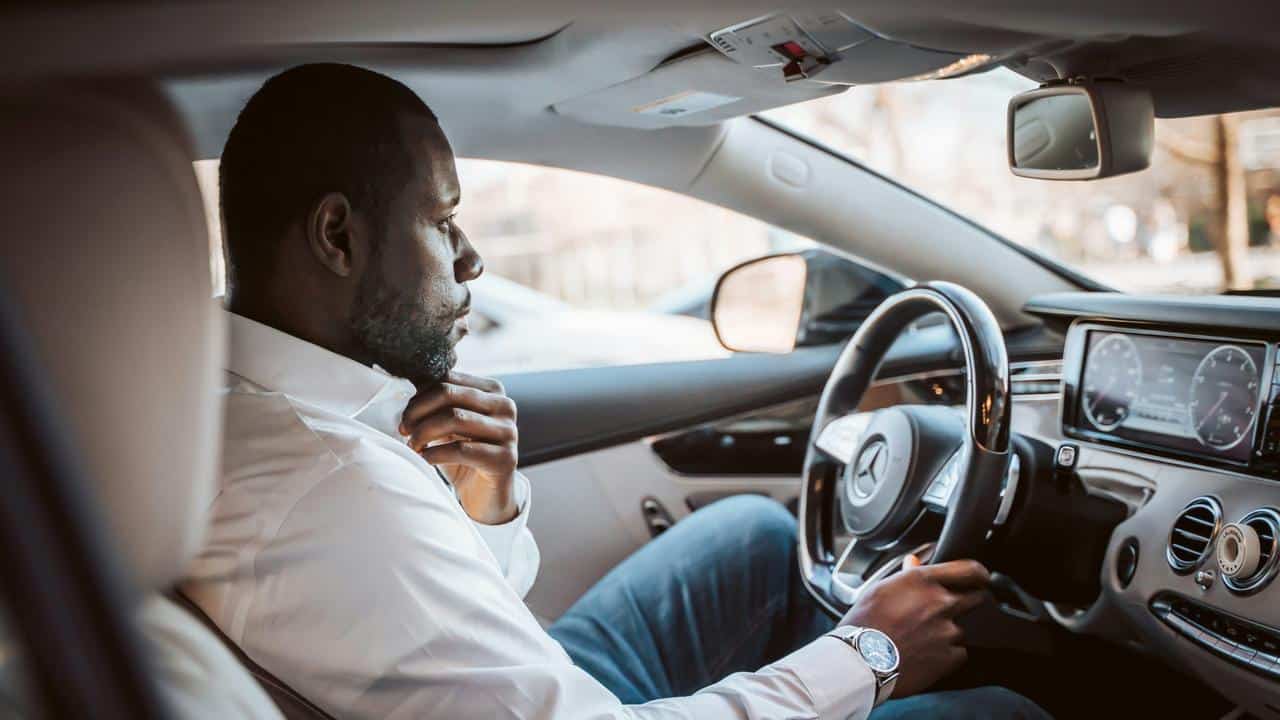 A man sitting in a car, holding the steering wheel and adjusting his collar.