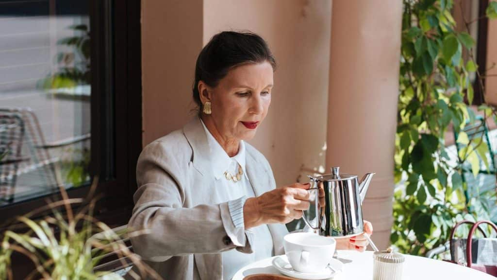 A woman in a light blazer pours tea from a silver pot into a cup at an outdoor café.