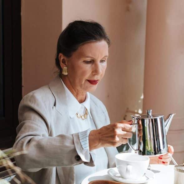A woman in a light blazer pours tea from a silver pot into a cup at an outdoor café.