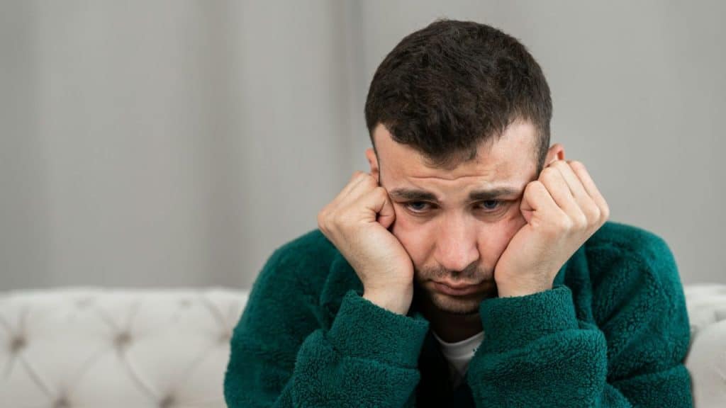 A distraught man rests his head in his hands, looking down with a sad expression.