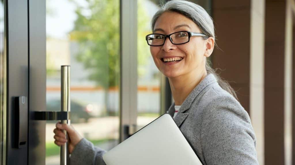 A smiling woman with graying hair and glasses opens a glass door while holding a white laptop.