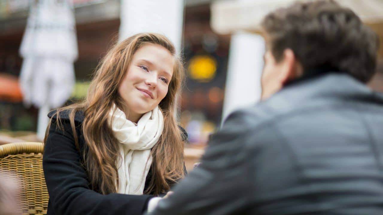 A young woman with long hair and a white scarf smiles at a man across a table outdoors.