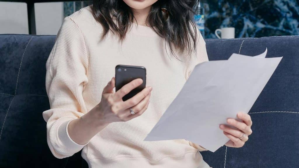 A woman sitting on a couch holding papers while looking at her phone.