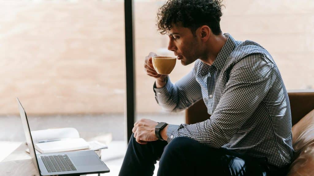 A man drinking coffee while working on a laptop in a bright café.