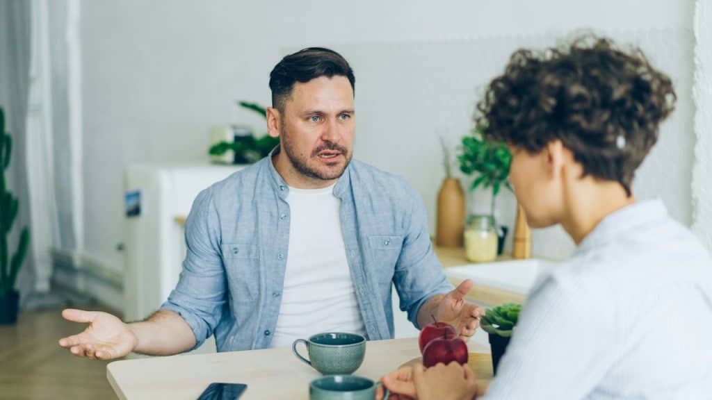 A man is sitting at a kitchen table talking to a woman, gesturing with his hands during a discussion.