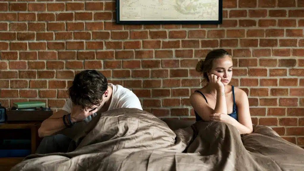 Couple sitting in bed with a duvet pulled up, both looking unhappy and avoiding eye contact after an argument.