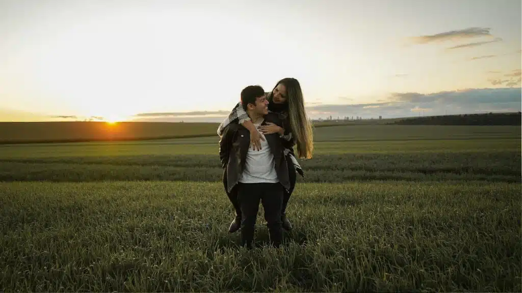 A woman riding on a man’s back in a green field during sunset.