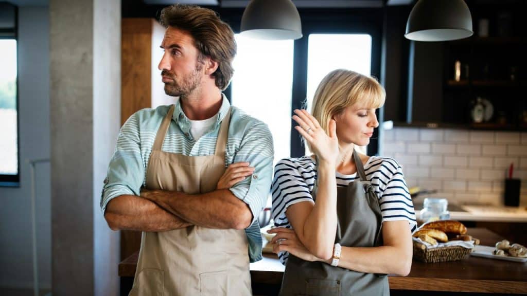 A frustrated couple wearing aprons stands back-to-back in a kitchen.