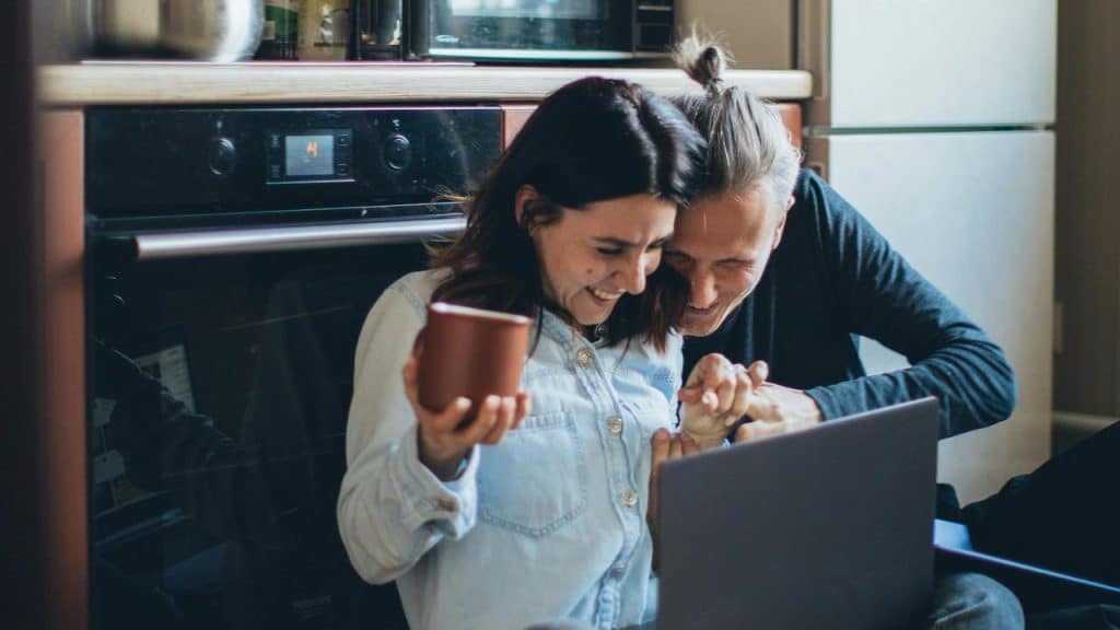 A couple sitting on the kitchen floor laughing over a laptop.