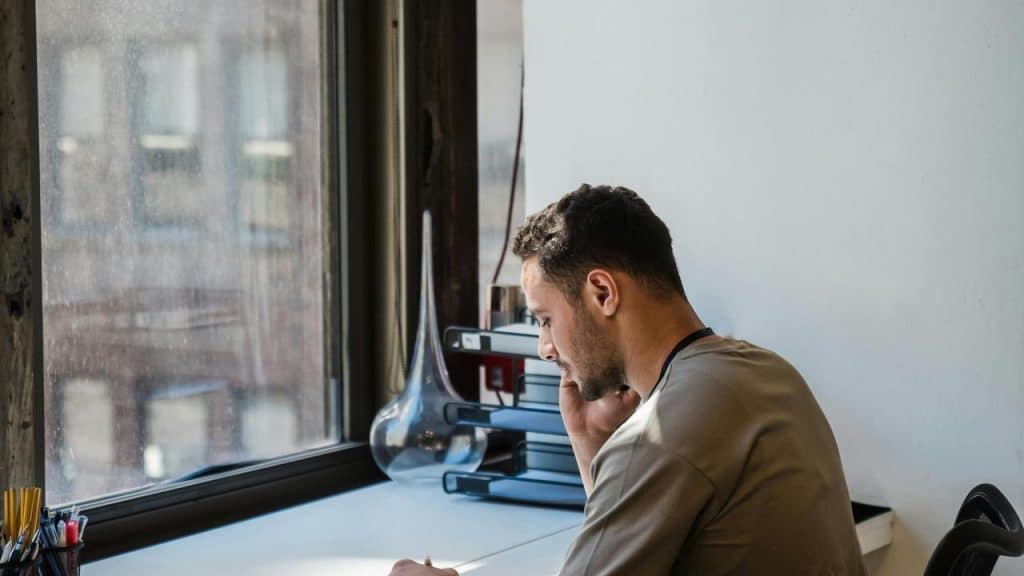 A man sitting at a desk by a window while talking on the phone.
