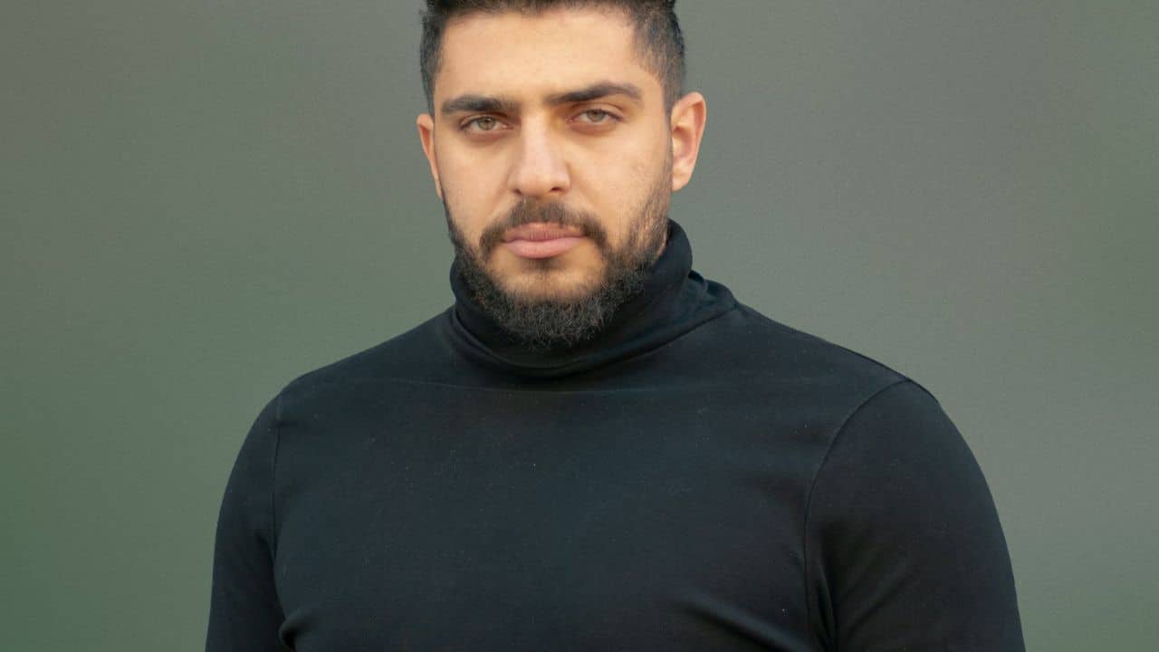A man with a beard and dark hair wearing a black turtleneck looks directly at the camera.