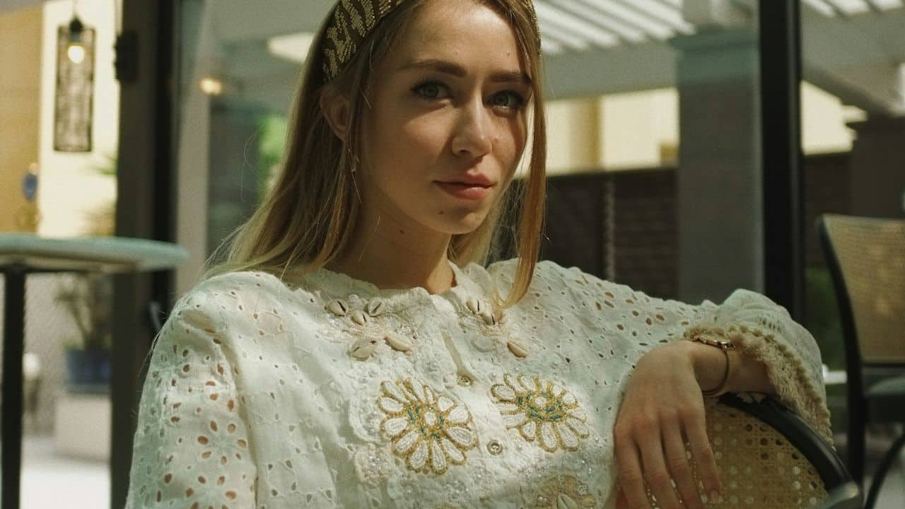 A young woman with long blonde hair and a headband wears an embroidered white jacket.