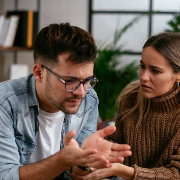 A man and woman sit on a couch, talking intensely and gesturing with hands.