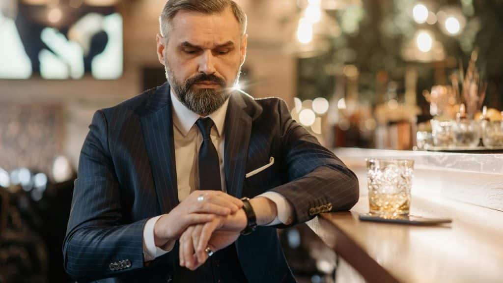 A man in a suit sitting at a bar and looking at his watch.