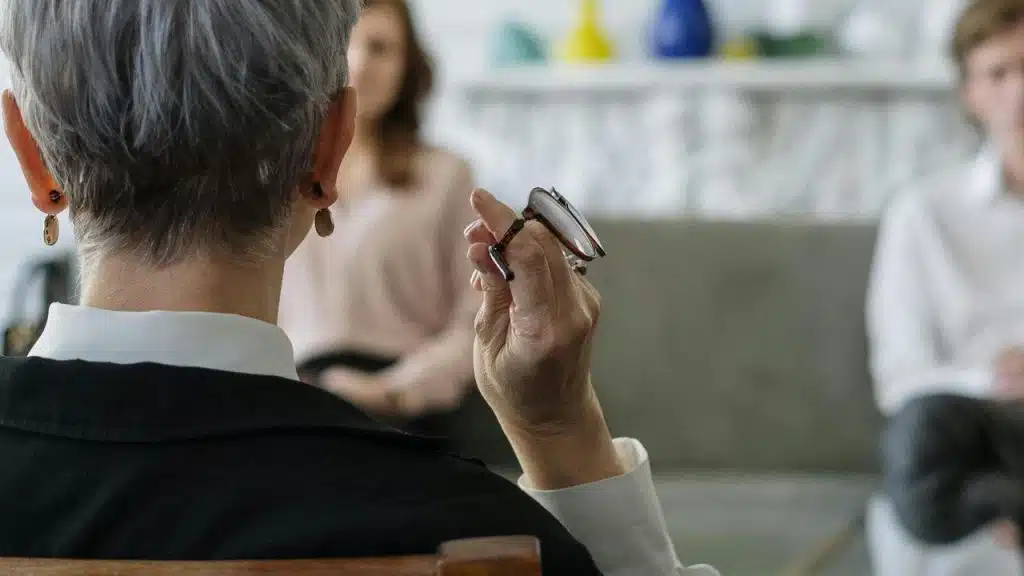 An older person holding glasses while speaking to two people seated in front of them.