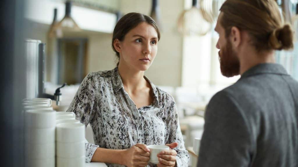 A woman holding a white mug is engaged in a serious conversation with a man indoors.