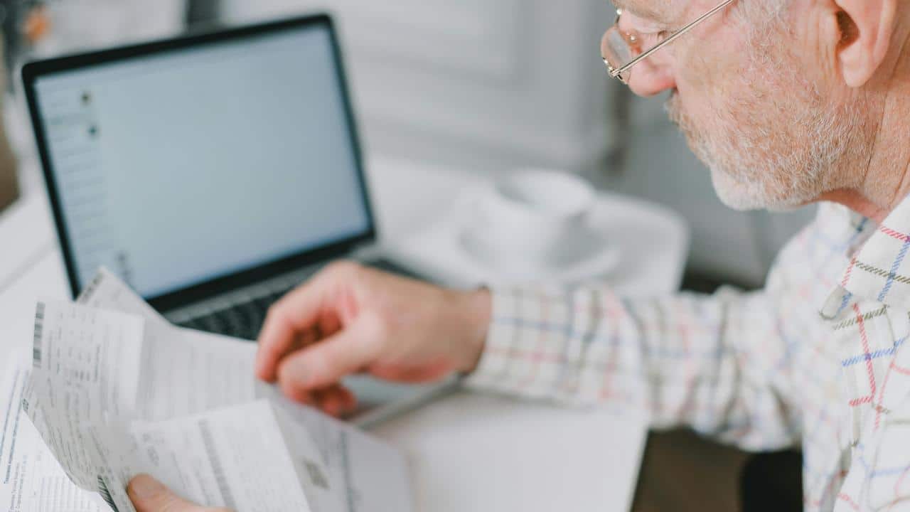 A senior man reviewing papers beside a laptop.