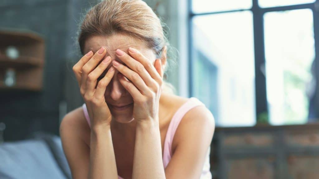 Blonde woman with slicked-back hair covering her face with both hands.