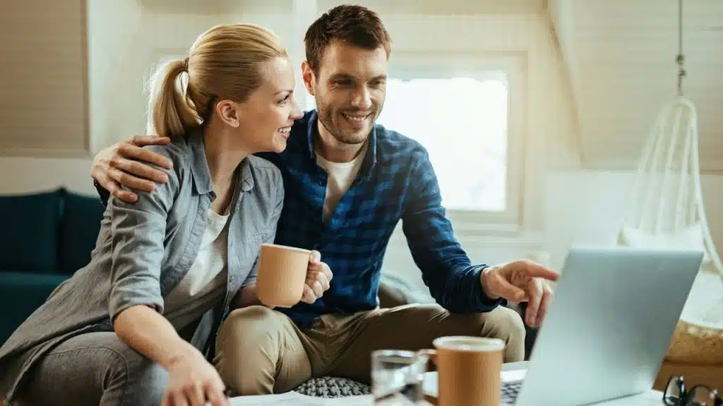 Smiling couple sitting close, looking at a laptop and holding coffee mugs.