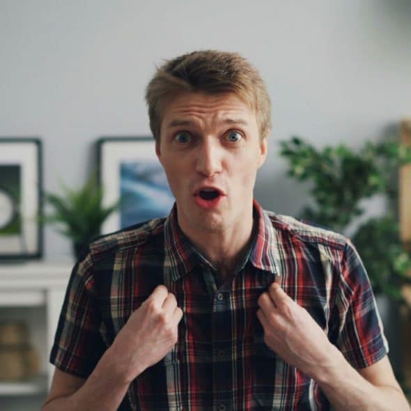 A man with a surprised expression and open mouth is holding his plaid shirt collar.