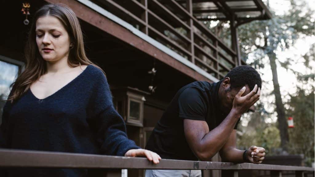 A distressed man with his head in his hands and a sad woman stand on a balcony.