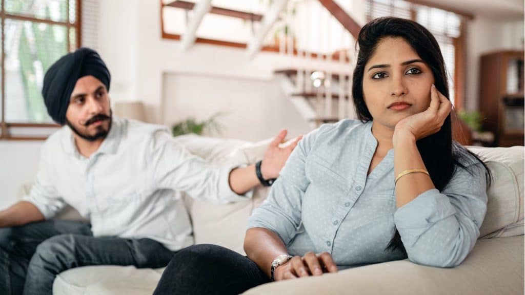 A woman is sitting on a couch with a distant expression while a man in a turban sits behind her, gesturing toward her.