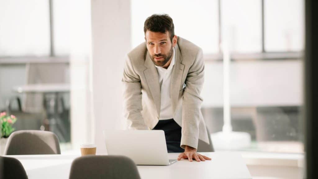 A man in a blazer is leaning over a laptop on a white conference table.
