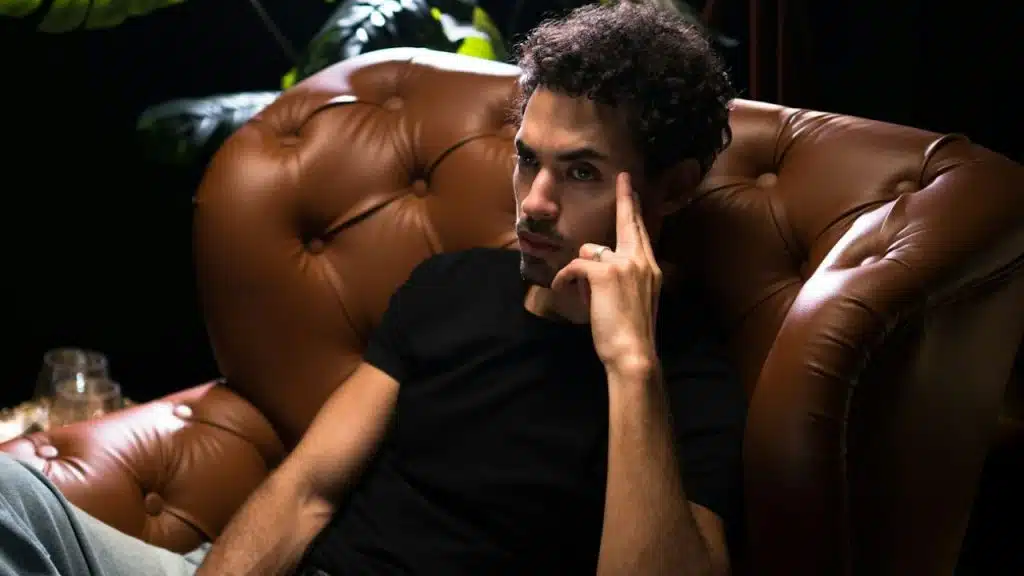Pensive man with dark, curly hair sitting in a brown leather armchair.