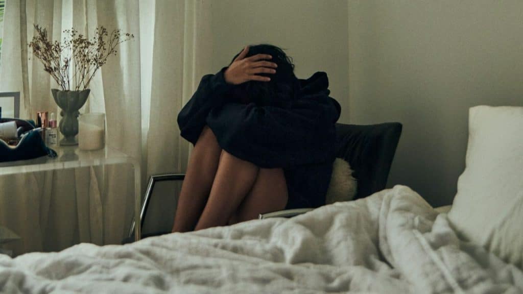 Woman sitting and hugging her knees, head covered, next to a bed.