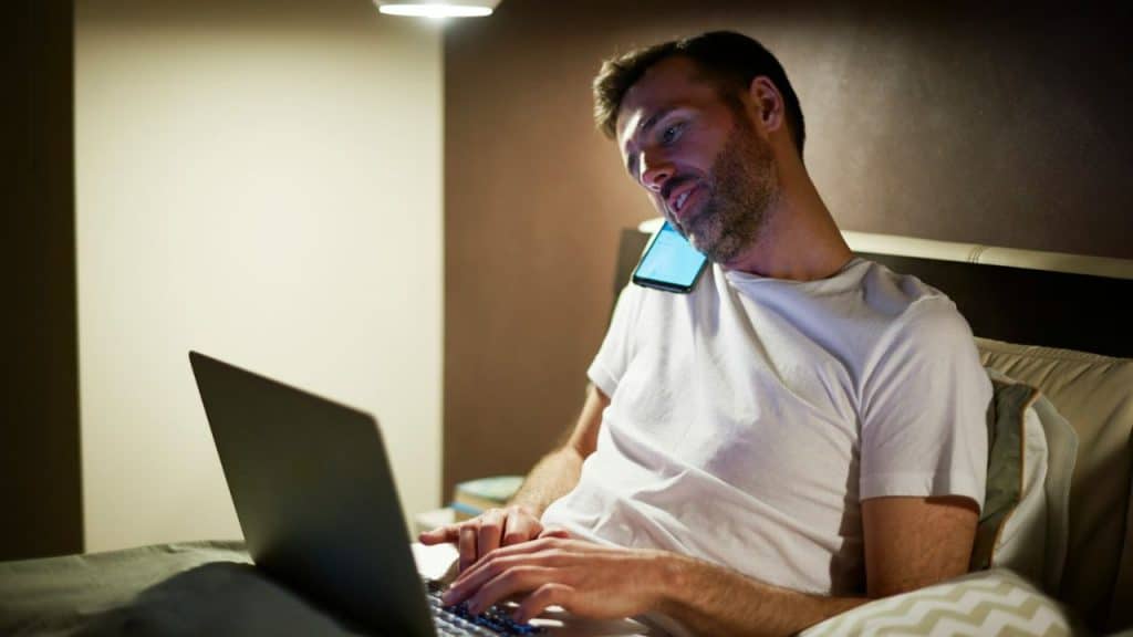 Man on a bed working on a laptop while holding a phone between his shoulder and head at night.
