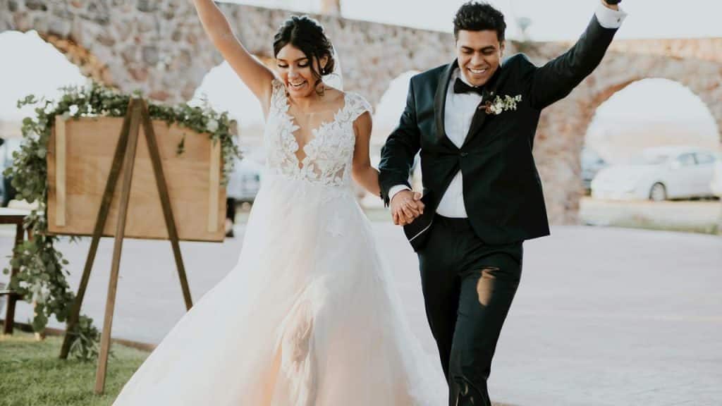 A joyful bride and groom hold hands and cheer while walking together outdoors.