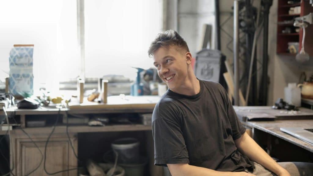A smiling man sitting in a workshop near a workbench.