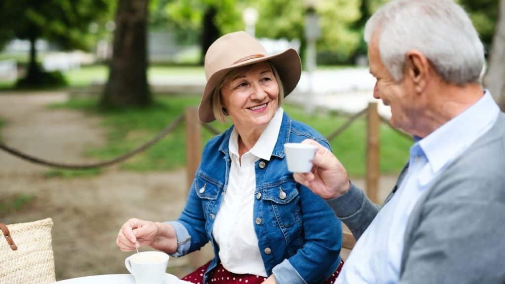 A smiling woman in a hat and denim jacket shares coffee with a man outdoors.