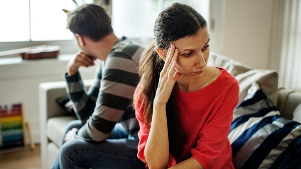 A sad woman rests her head in her hand while sitting on a couch, with a man turned away in the background.
