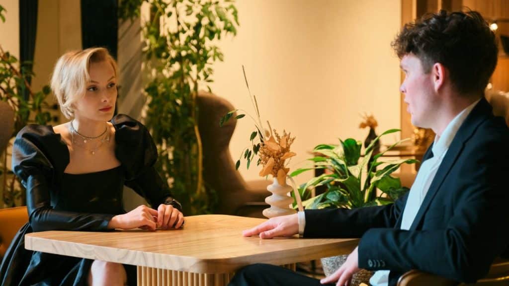Two people, one in a black dress and one in a suit, talking at a wooden table.